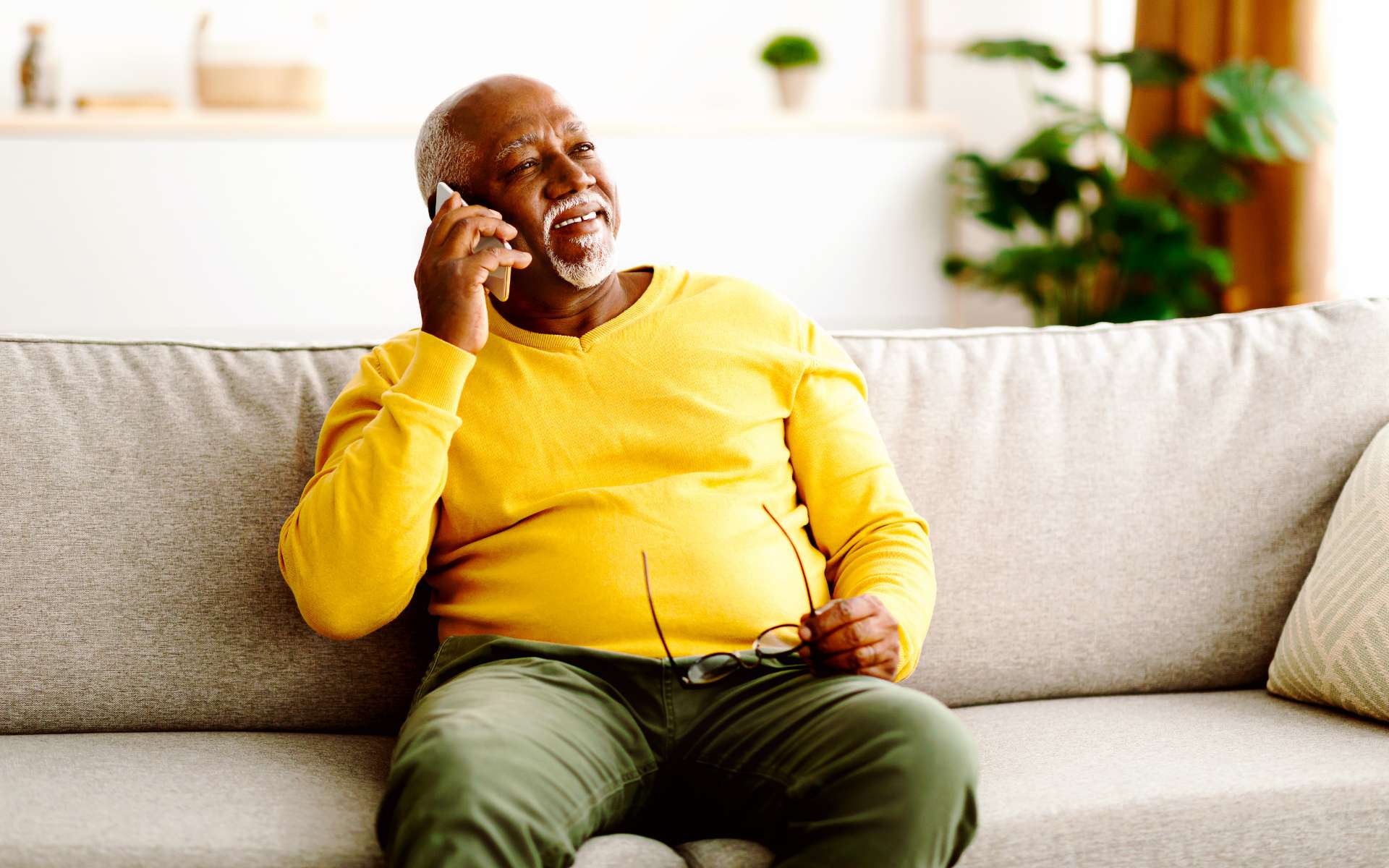 An older, dark-skinned man sitting on a comfortable chair, talking on the phone. 