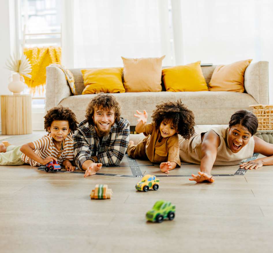 A black woman, a man with medium skin tone, and their two children are all smiling, playing on the floor of a bight, sunny living room.