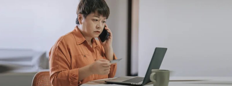 A middle-aged Asian woman with short hair stares at her laptop screen, frowning slightly with a worried expression. She sits at a table, holding her credit card in one hand, while pressing her phone to her ear with the other.
