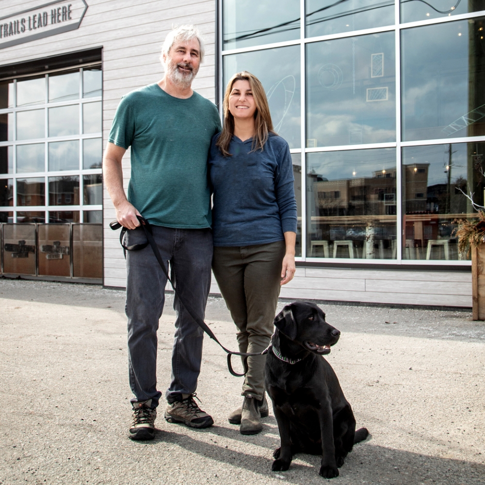Meridian Members, Rob Garrard and Joanne Richter. Rob and Joanne have been Members since 2014. They are outside, near a store front, with their black dog on a leash.