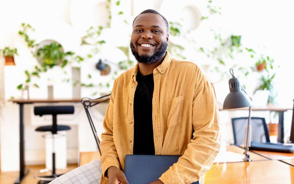 Smiling black man leans against a deck in a bright, plant-filled work space.