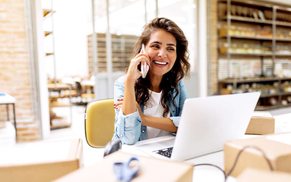 Smiling young woman with medium skin tone and long dark hair is on the phone, sitting in front of a laptop in a sunny warehouse.