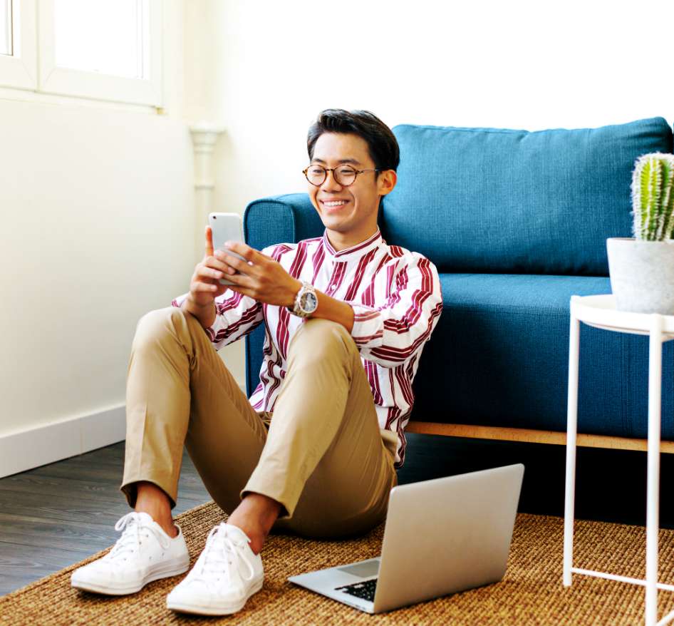 A young man with lighter skin, black hair, and glasses smiles at his phone, He is sitting on the floor, leaning against his couch.