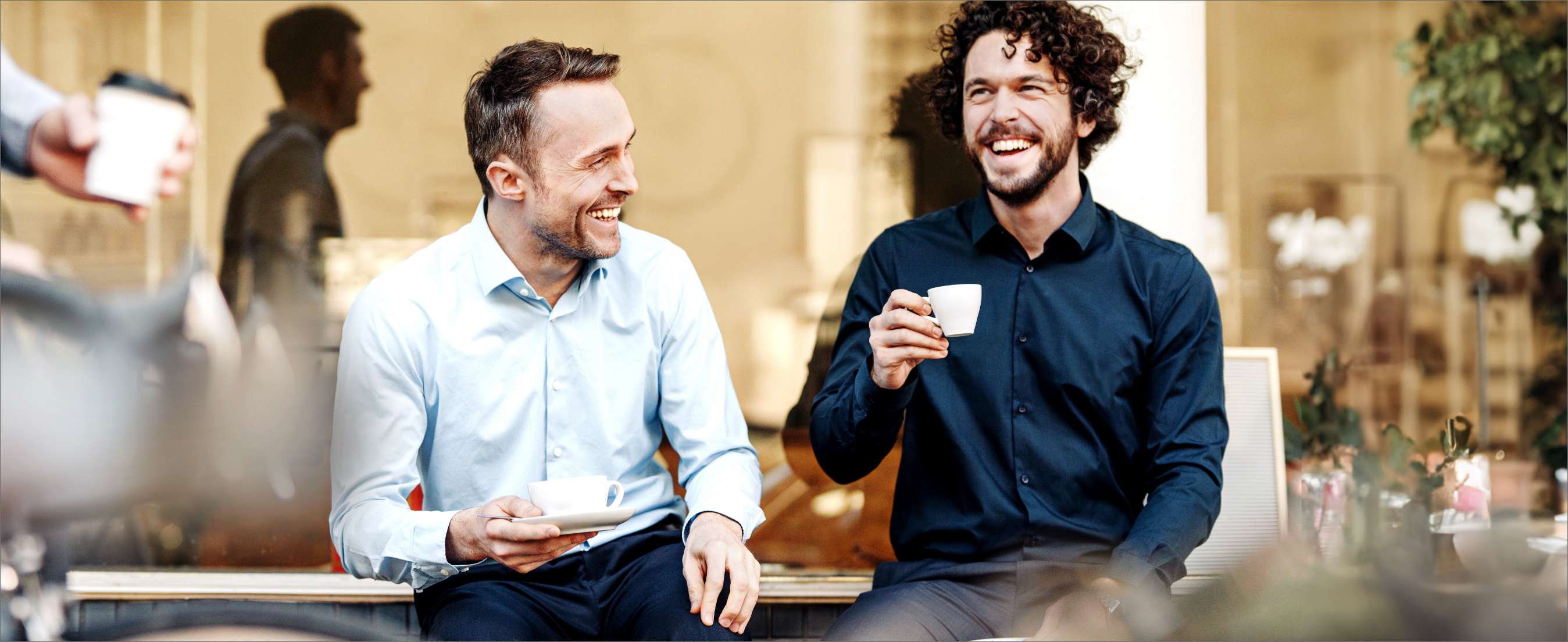 Two white men laughing outside a café, holding espresso cups.