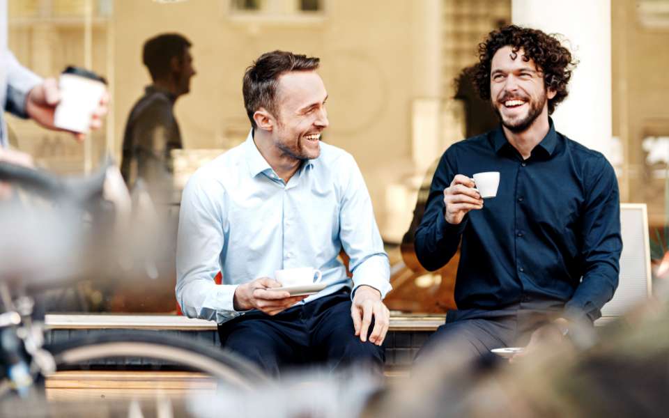 Two white men sitting in front of a coffee shop, laughing and drinking coffee