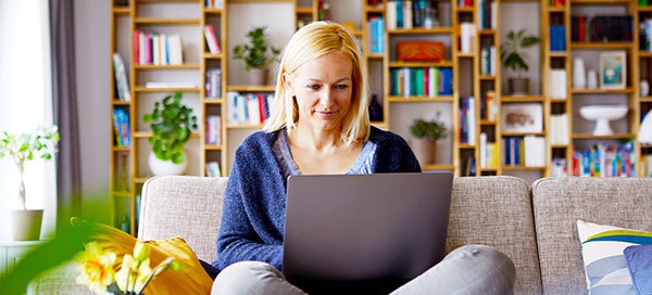 A woman with blond hair sits cross legged on her couch, smiling at her laptop.