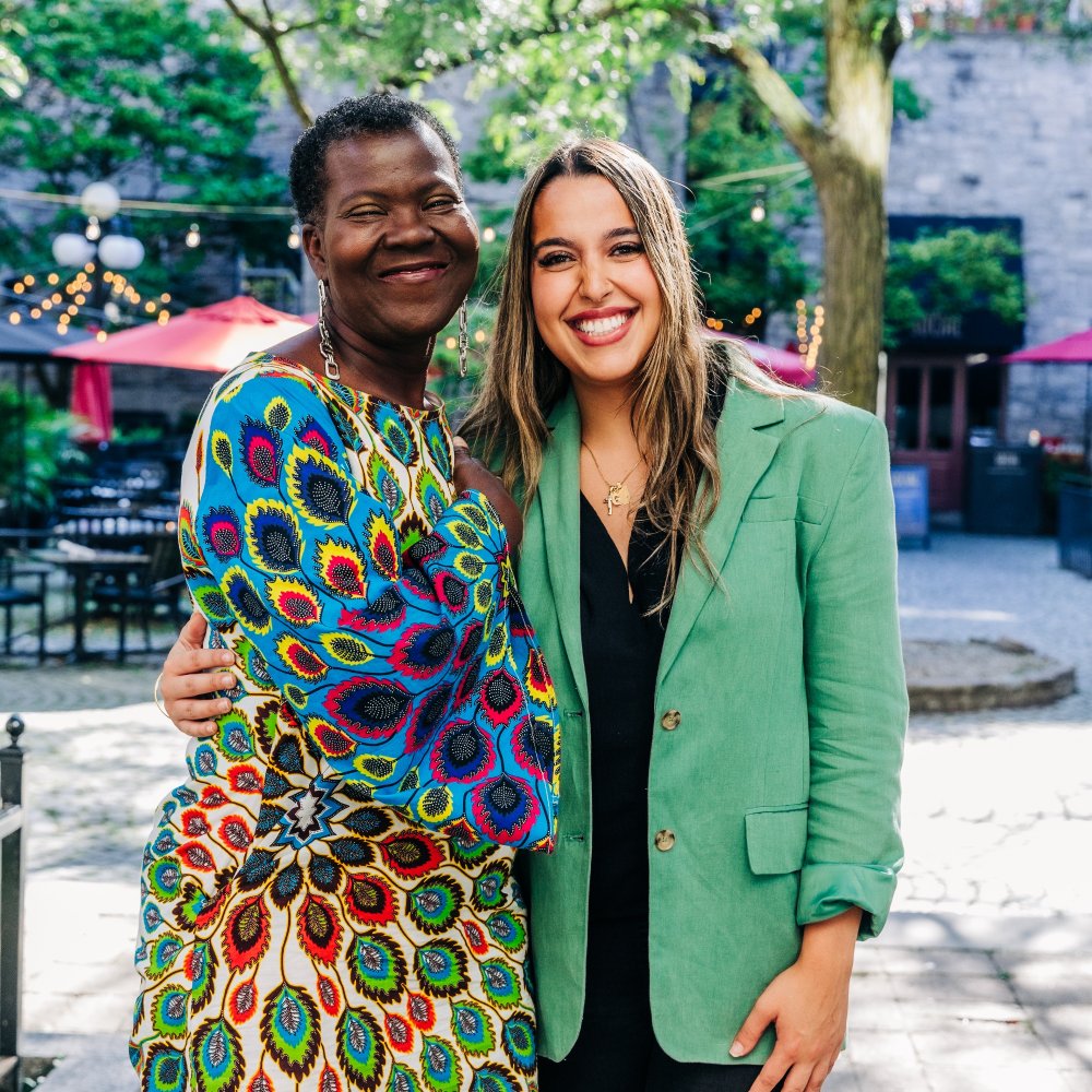 Two women stand outdoors smiling; one wears a colorful patterned dress, the other a green blazer. They are close together with one arm around each other. Trees and patio seating are in the background.