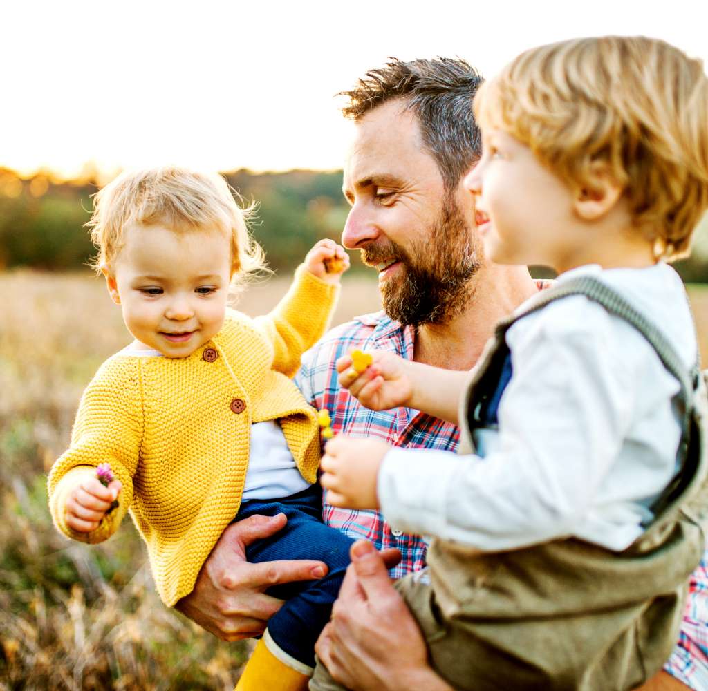 A father holding two toddler children on a meadow outdoors in autumn.