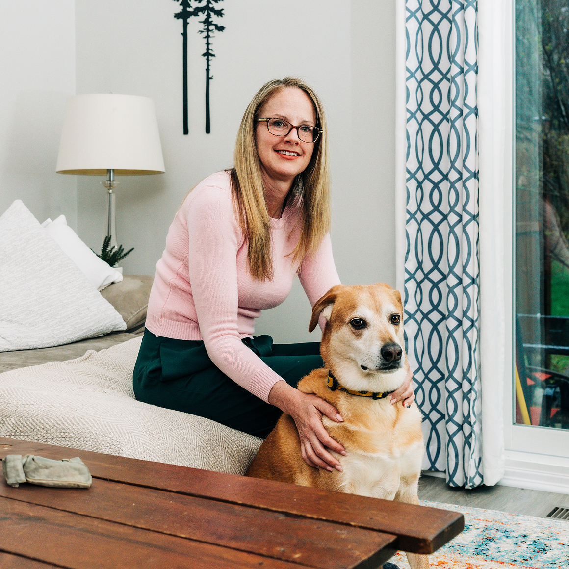 Light skinned woman with blond hair, sits with her dog at home.