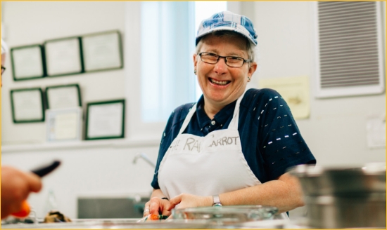 Raw carrot employee wearing an apron and smiling in the soup kitchen. She is older, with short grey hair and glasses.