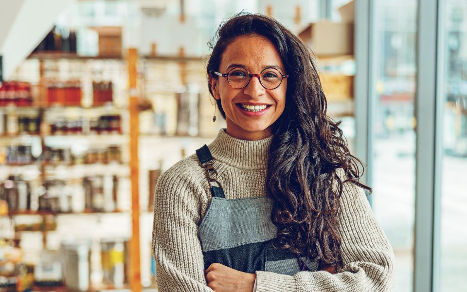 Smiling young woman with long dark curly hair, medium skin tone and glasses, smiles and crosses her arms. She is standing in a small retail store and wearing an apron.