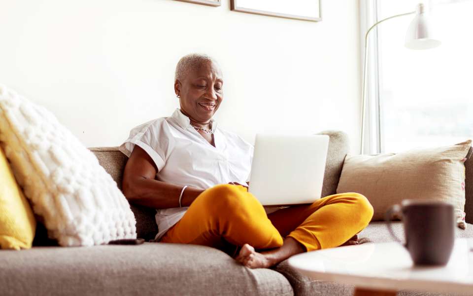 An older woman sits cross legged on her sofa with her laptop