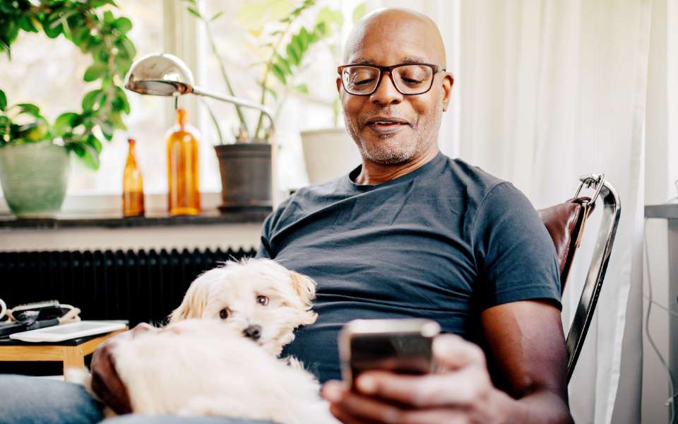 A man sits in a chair at home with a dog in his lap. He’s reading something on his mobile phone.