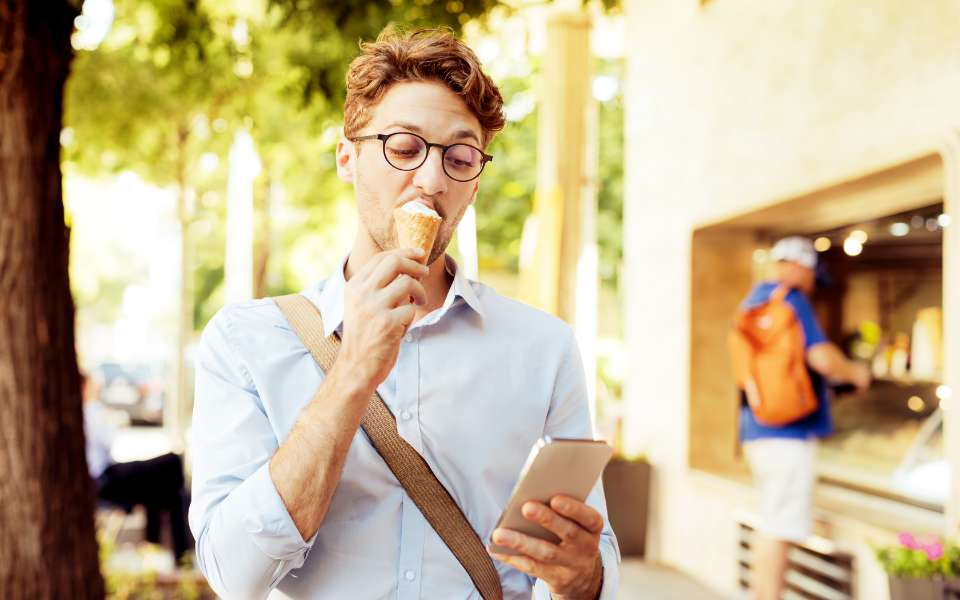 A male student walks outside, eating an ice cream while glancing at his mobile phone