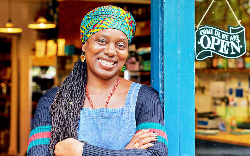 A women smiles, leaning against the door frame of her store. There is a sign in the window that says “we are open.”