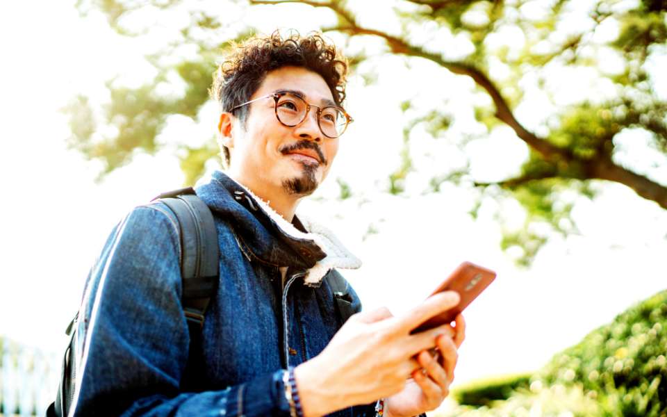 A man smiles, holding his phone. Trees and parkland in the distance.