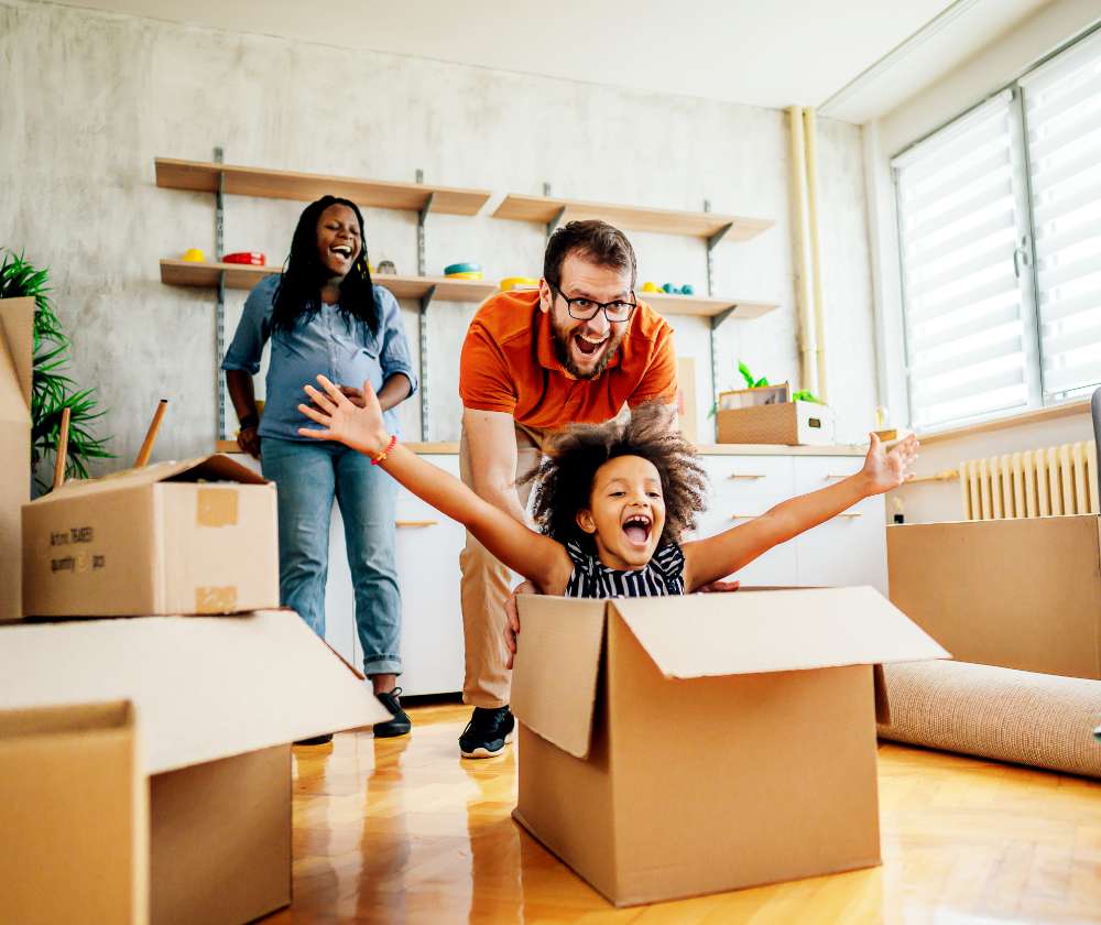 A black pregnant woman laughs and smiles while her husband pushes their child in a moving box. They are unpacking their living room. 