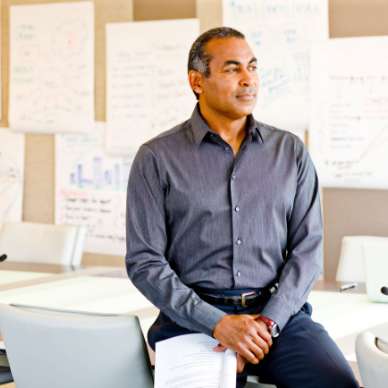 A man with medium skin tone and greying hair leans on a desk in a bright workspace filled with charts.