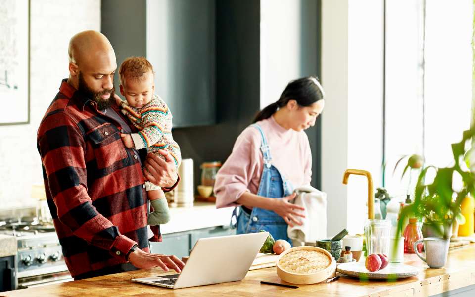 A middle aged Black man with beard is standing at a kitchen island. He is holding a baby and looking at a laptop. A middle aged Asian woman is standing beside him, washing dishes. 