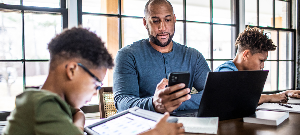 Middle age man using a smartphone while working on a laptop at a table with two children using digital devices nearby in a bright home setting.