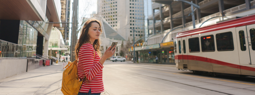 A woman with a yellow backpack and striped shirt stands on a sidewalk looking at her phone with tall buildings and a light rail train in the background.