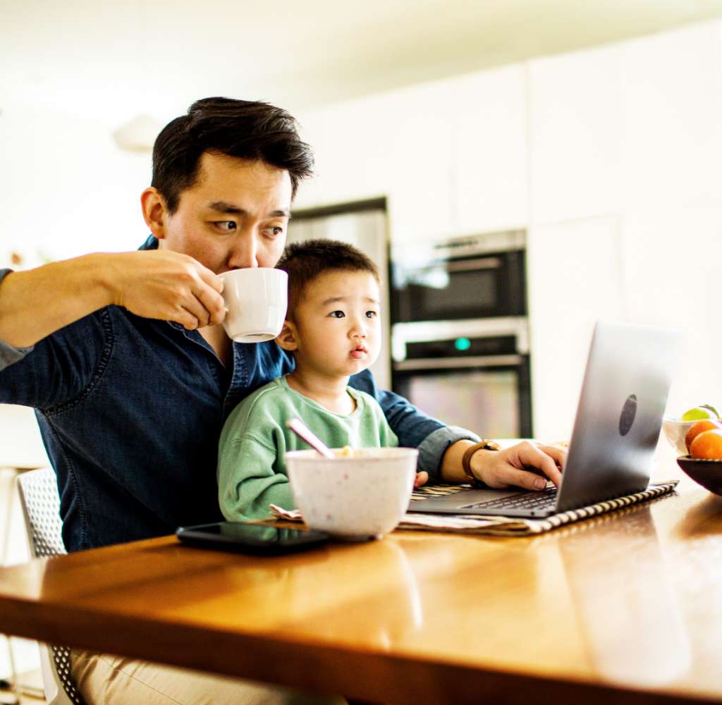 Mid adult Asian man sipping out of a coffee mug with his young son sitting on his lap, at the kitchen table. He is looking at his laptop.
