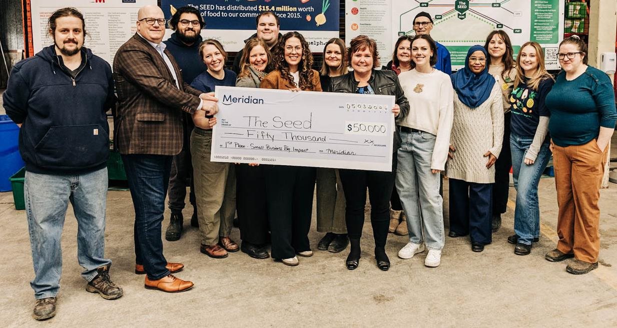 Inside a warehouse with signs about The SEED, a group of smiling people hold up a cheque for $50,000.