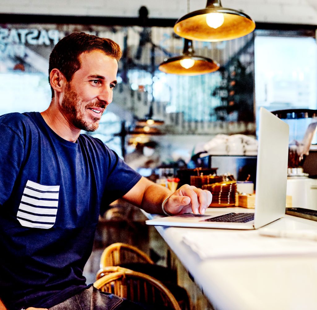 A smiling bearded man works on a laptop at the counter of a bright cafe.