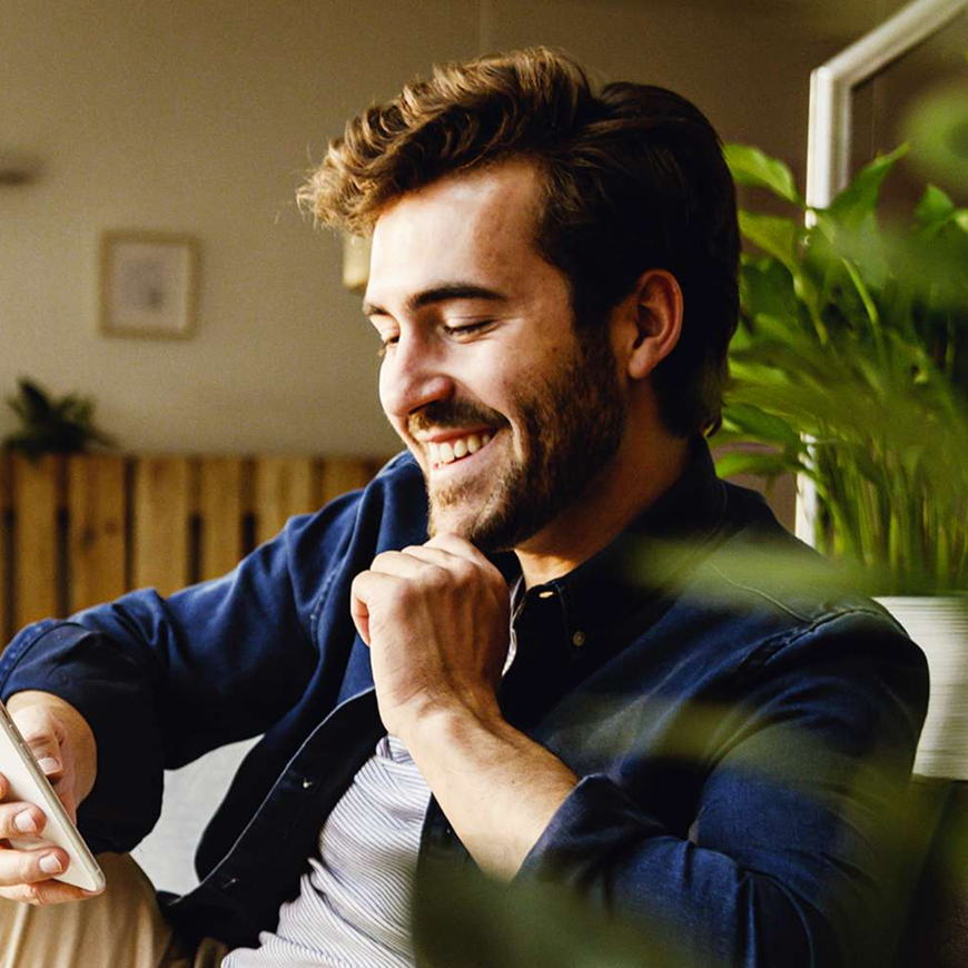 A man sitting on a couch smiling as he uses his phone