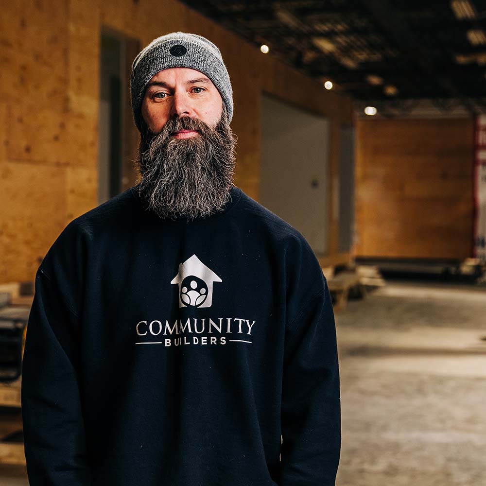 Light skinned man with a full beard, wearing a cap and community builders sweatshirt. 
