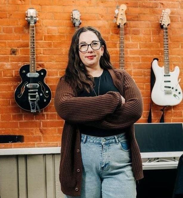 A woman with long dark hair and glasses stands in front a set of electric guitars hanging on a wall.