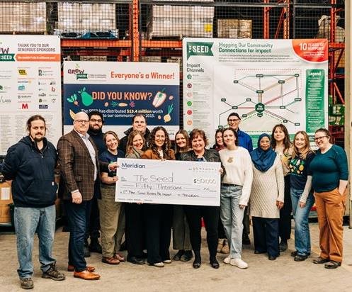 Inside a warehouse with signs about The SEED, a group of smiling people hold up a cheque for $50,000.