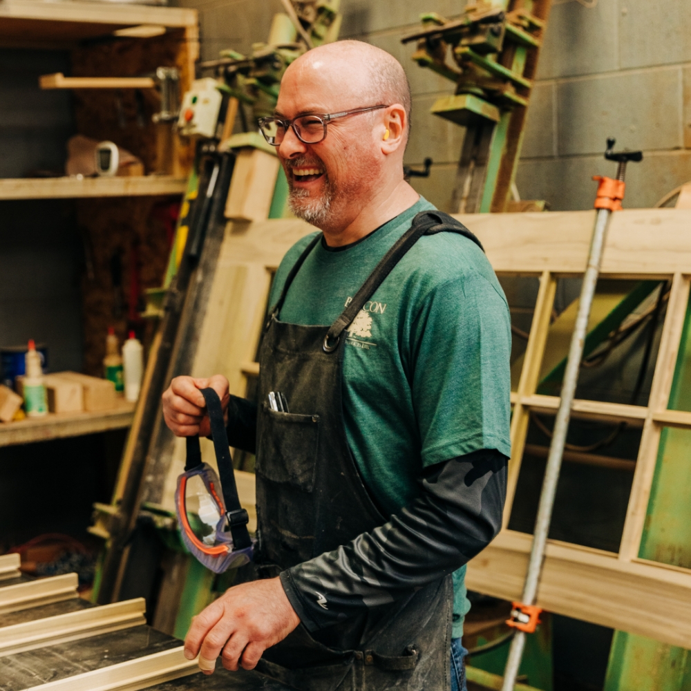 Employee at Byrcon Wood Products is in the workshop, wearing overalls and holding a pair of protection glasses. 
