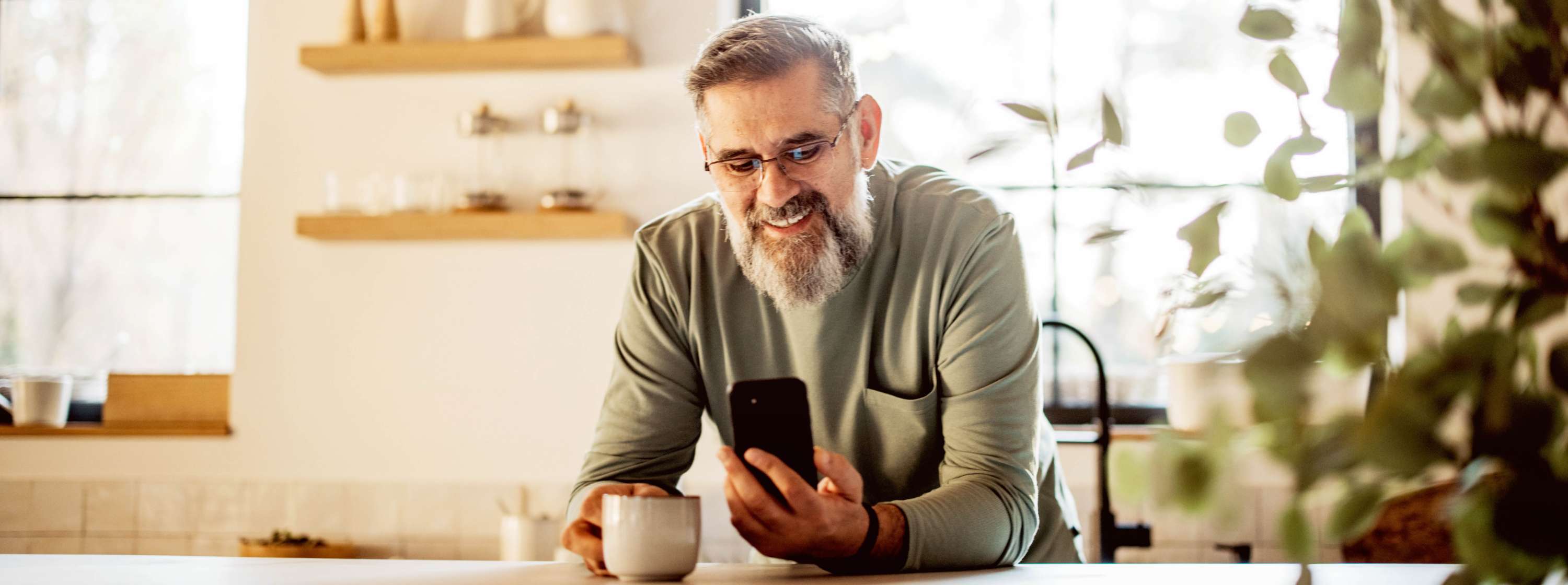 A middle-aged man with grey hair and long scruffy beard, sitting at a table, holding a smartphone in one hand and a cup in the other in a kitchen.