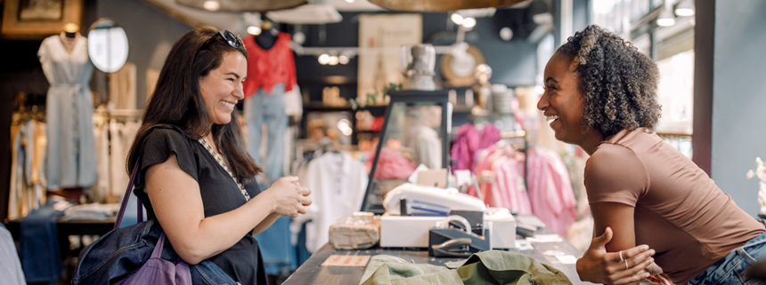 Two women smiling and talking at a clothing store counter with clothes in the background.
