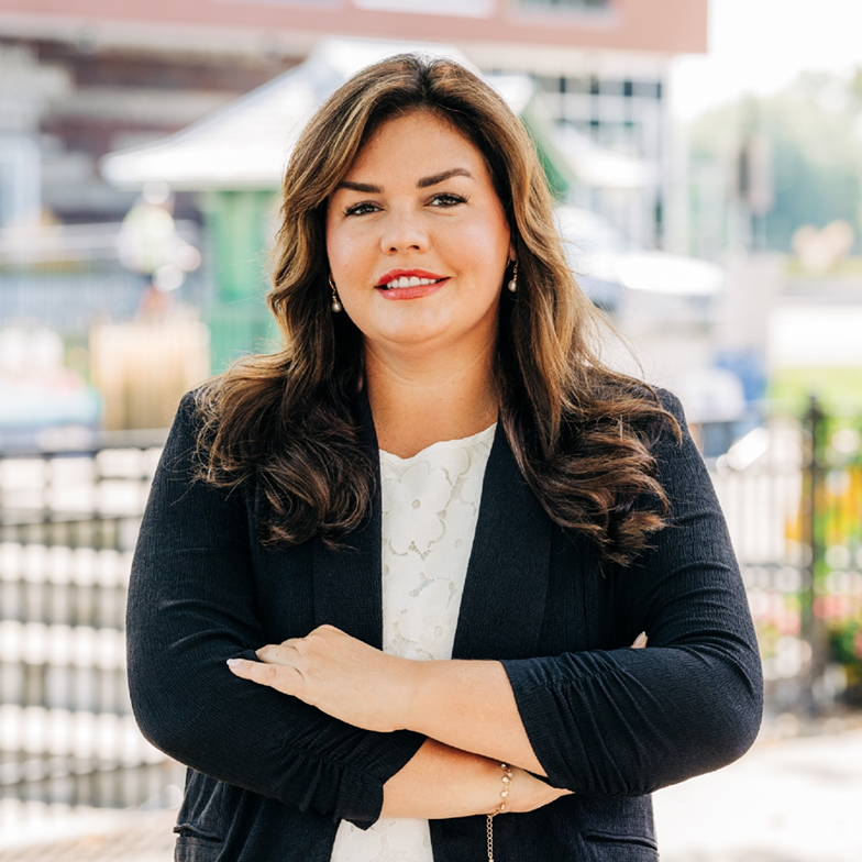 A confident woman with brown hair stands outdoors, arms crossed, wearing a dark blazer over a white top. The background shows blurred urban scenery.