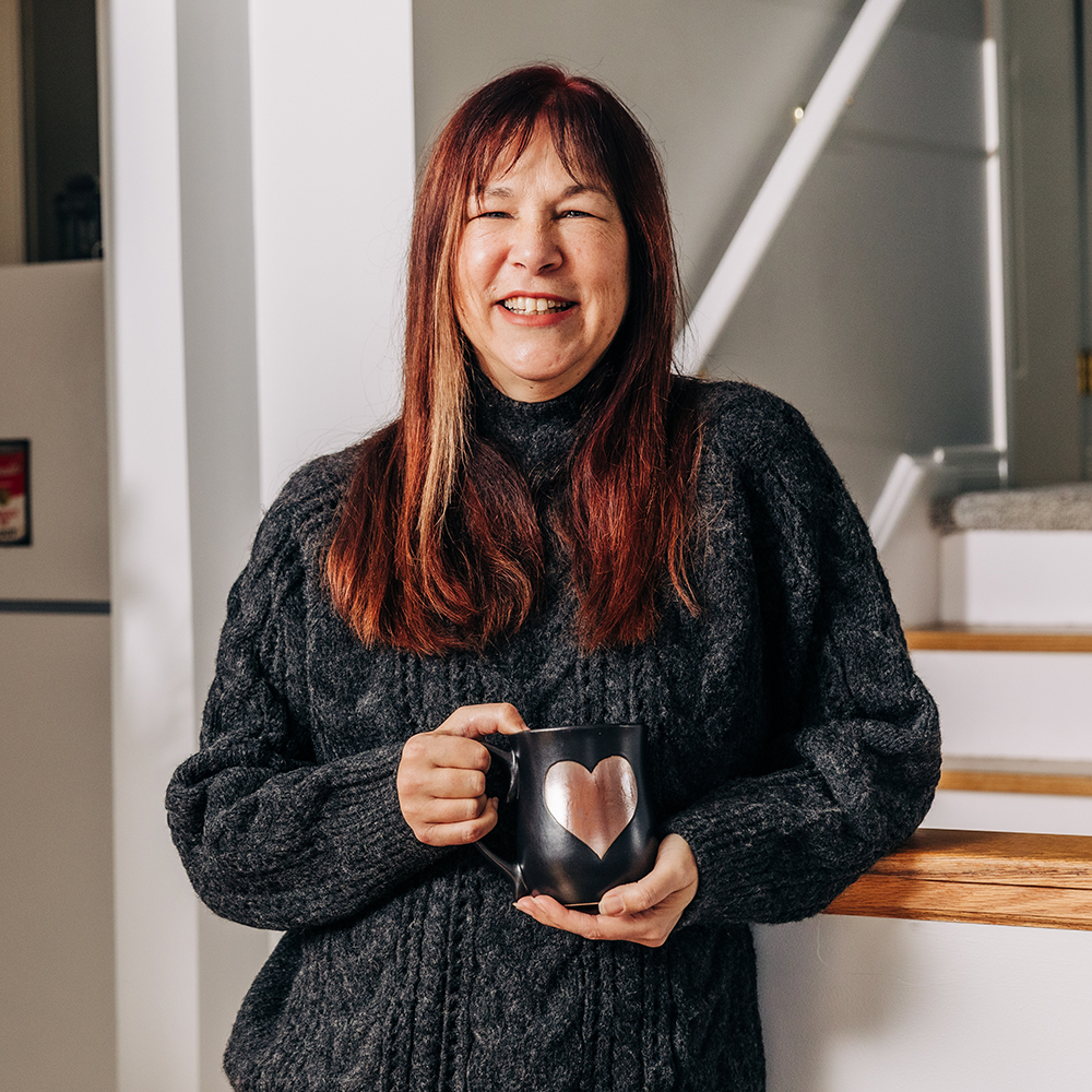 A smiling woman standing, holding a large cup of black coffee and wearing a wool sweater.