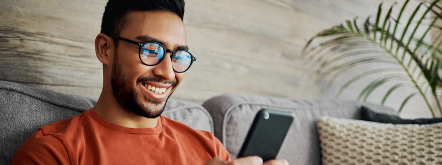 A young man in an orange shirt and glasses sits on a gray couch smiling at their smartphone