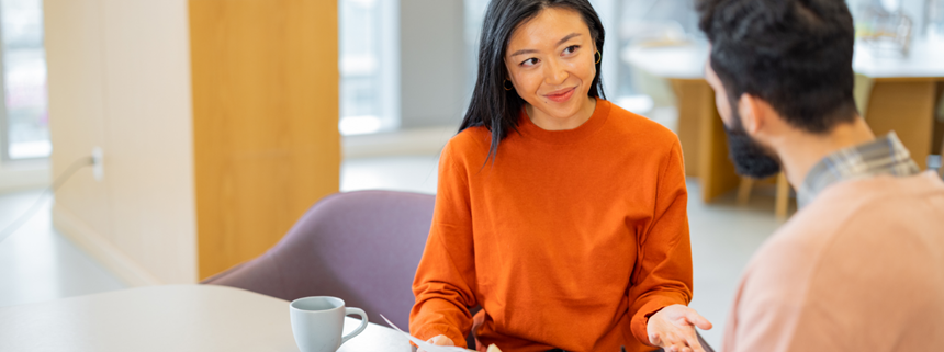 A young woman in an orange sweater talks with a colleague at a table with a laptop and mug.