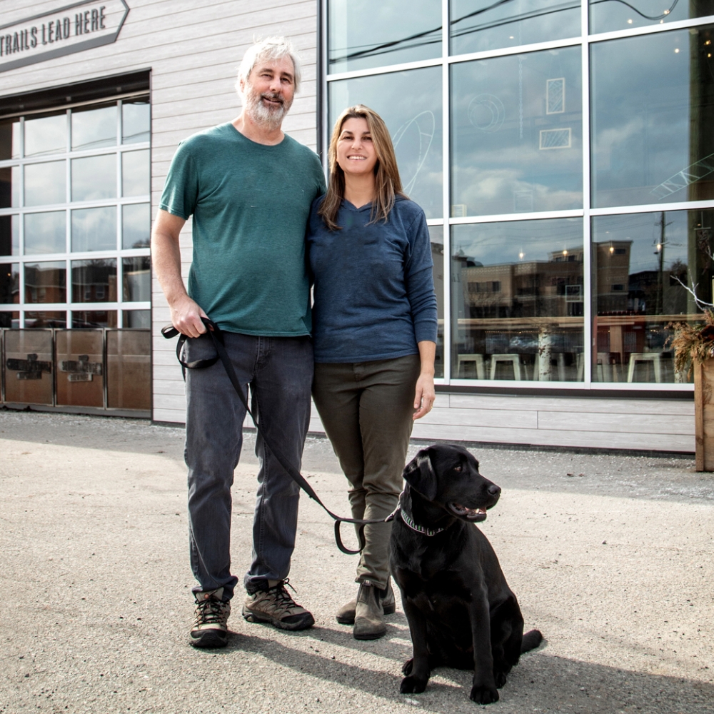 Meridian Members, Rob Garrard and Joanne Richter. Rob and Joanne have been Members since 2014. They are outside, near a store front, with their black dog on a leash.  