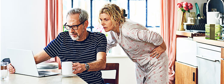 A white middle-aged man and woman wearing pajamas are in their kitchen looking at a laptop. The man is sitting at the table holding a mug and the woman is leaning over his shoulder.