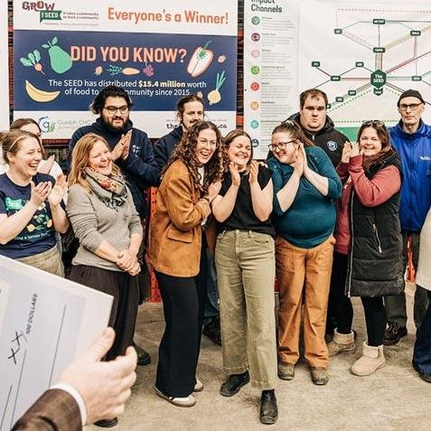 Inside a warehouse with signs about The SEED, a group of smiling people are clapping and hugging each other. 