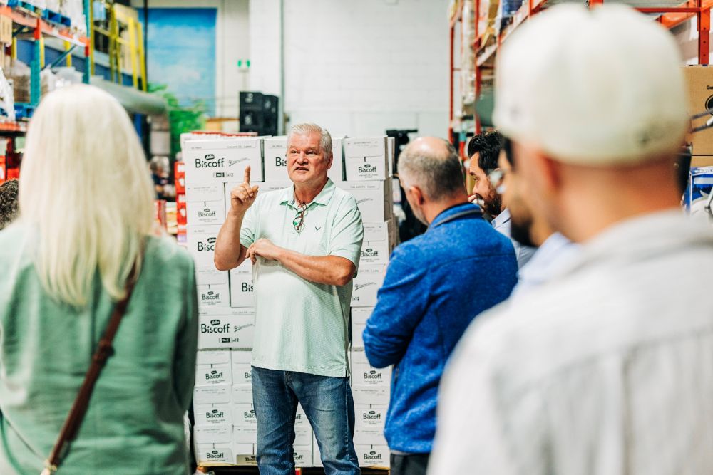 A warehouse guide speaks to a group of visitors gathered between storage shelves, with stacks of product boxes visible behind the speaker.