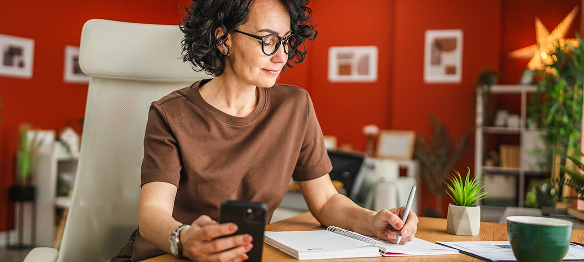 A middle-aged white woman sitting at desk writing in notebook and holding phone