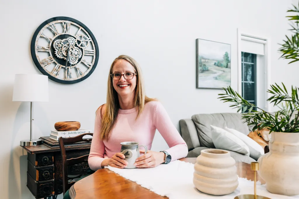 Stacy M - Meridian Member since 2021 A smiling woman sits at a dining table holding a hot beverage mug surrounded by home furnishings that include a clock, a plant, and a couch.