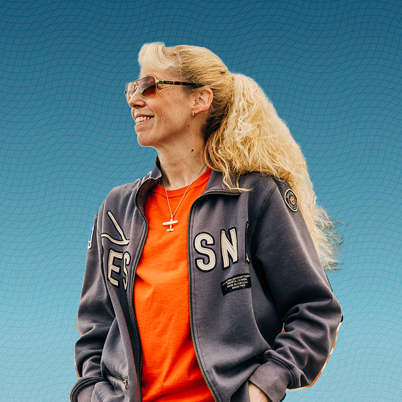 Woman with long blonde hair and sunglasses, wearing a zip-up sweatshirt and a bright orange t-shirt.