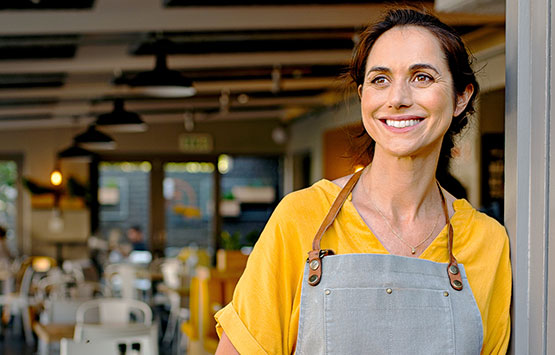 Business owner smiles as she stands outside of her restaurant, wearing an apron and holding a tablet