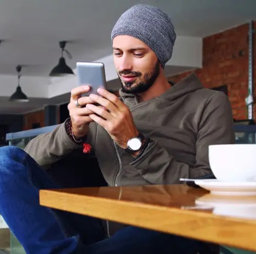 A young man with medium skin tone and a tidy beard sits in a bright coffee shop. He holds his phone with both hands, smiling slightly as he types.