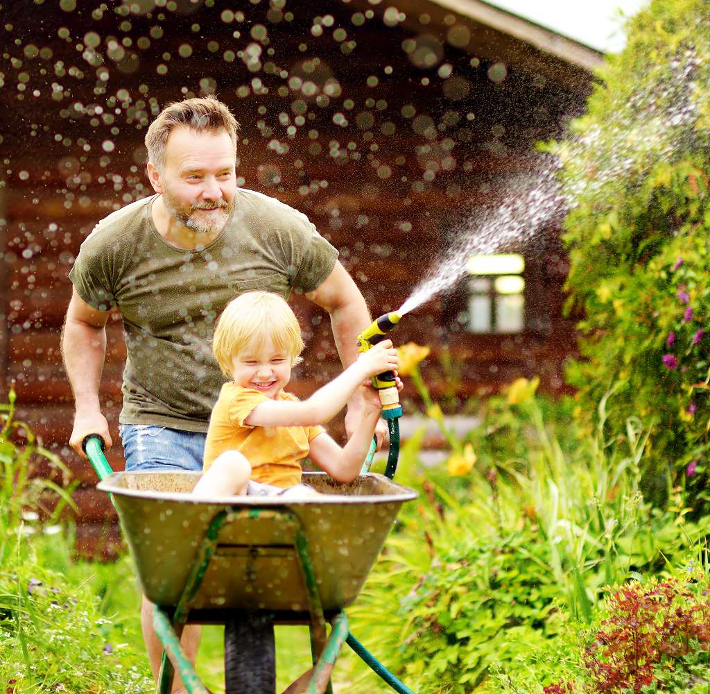 Middle aged white man pushes a laughing little boy with blonde hair in a wheelbarrow. The little boy is playing with a hose.