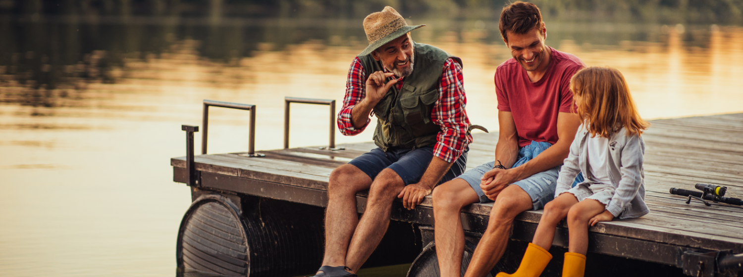 An older man wearing fishing gear, his son and his granddaughter sit on a dock at a lake during sunset, smiling and laughing together.